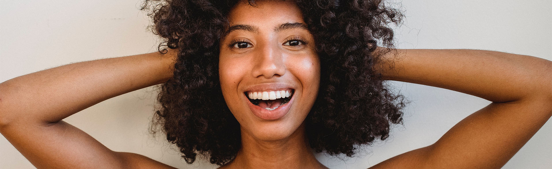 Lachende Frau mit lockigen Haaren hält Hände hinter dem Kopf.